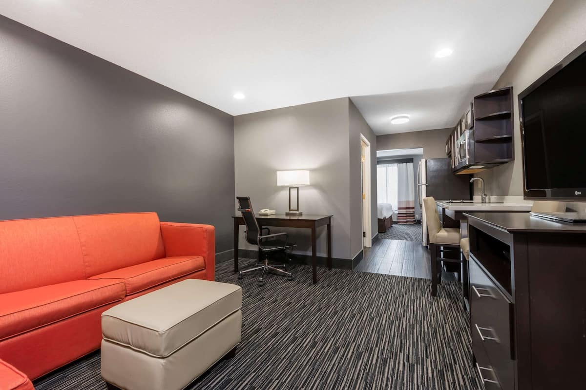 A hotel room with a sofa, a desk, a chair, a TV, and a kitchenette.