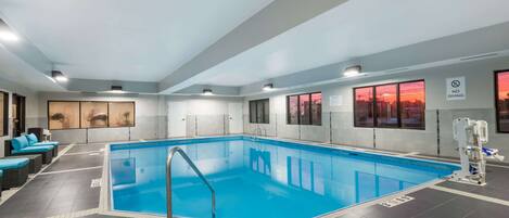 An indoor swimming pool with a clear view of the sunset through large windows, a lifeguard stand, and a seating area with blue cushions.
