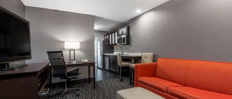 A hotel room with a flat-screen TV, a desk with a lamp, a kitchenette, and an orange sofa.