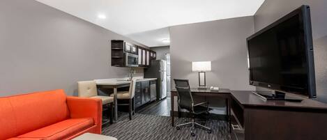 A hotel room with a sofa, a desk, a chair, a television, and a kitchenette.