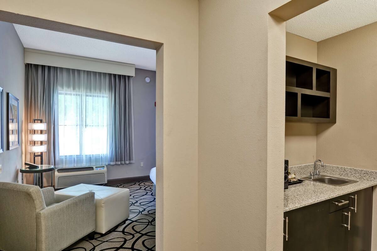 Habitación de hotel con sofá, una mesita de noche con lámparas, ventana con cortinas, kitchenette con fregadero y estante empotrado.