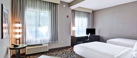 A hotel room with a bed, a desk, a chair, a lamp, and a window with curtains.