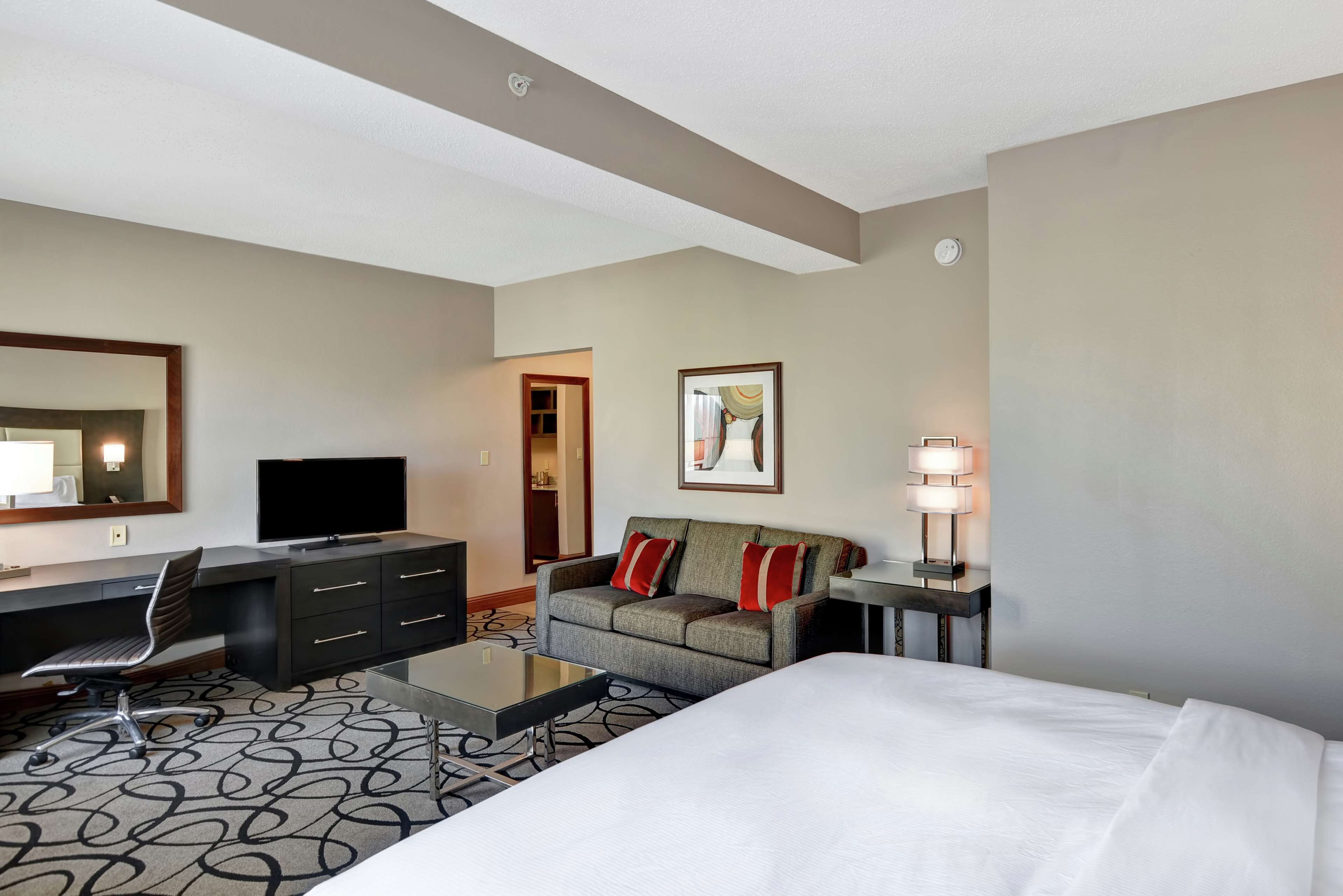 Ein Hotelzimmer mit einem Bett, einem Schreibtisch mit Stuhl, einem Fernseher, einer Couch mit roten Kissen, einer Lampe und einem Spiegel.