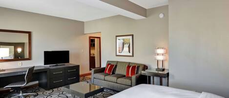 Ein Hotelzimmer mit einem Bett, einem Schreibtisch mit Stuhl, einem Fernseher, einer Couch mit roten Kissen, einer Lampe und einem Spiegel.