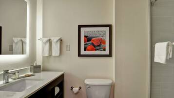 Une salle de bain avec un lavabo blanc, des toilettes, un tableau encadré au mur et une porte de douche en verre.