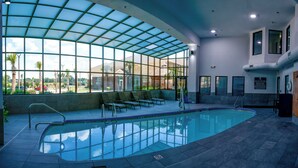 An indoor swimming pool with a glass ceiling, lounge chairs, and a view of the outside.
