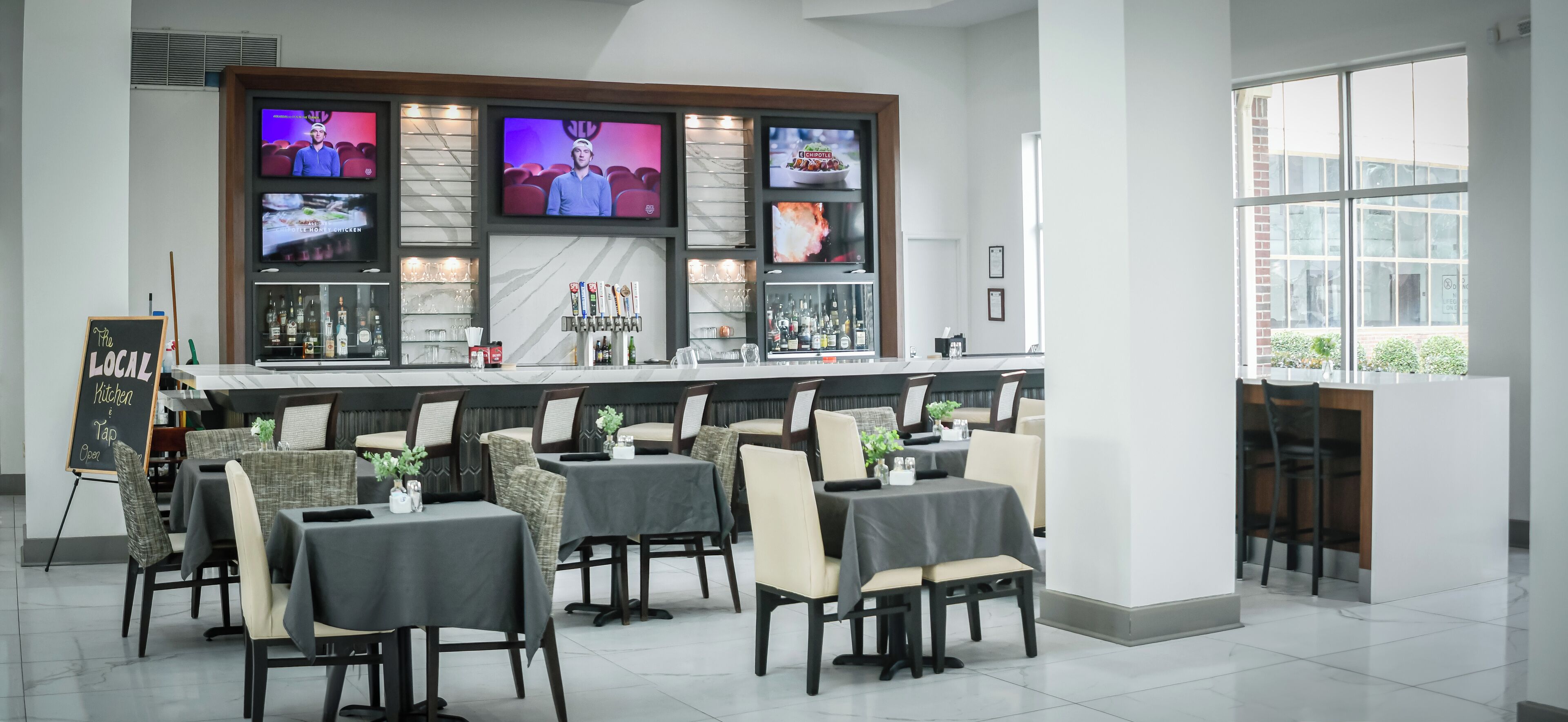 Un bar moderne doté de plusieurs écrans, d’un comptoir bien garni de boissons et de sièges confortables.