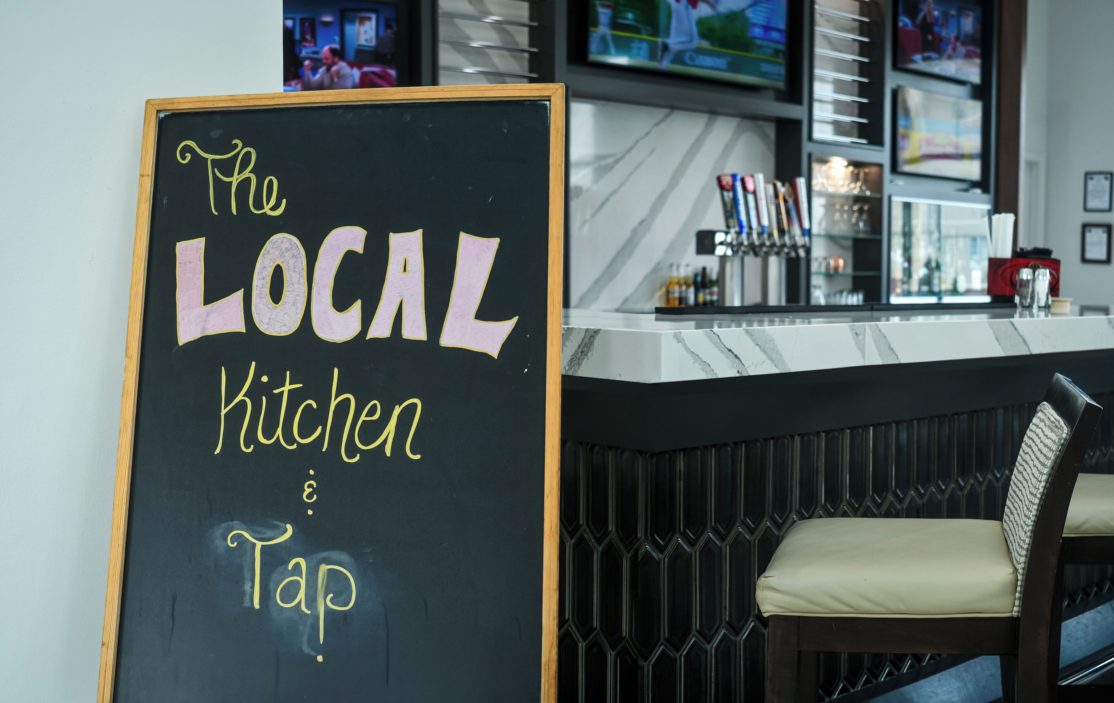 A chalkboard sign reading "The Local Kitchen Tap" in a modern bar setting.