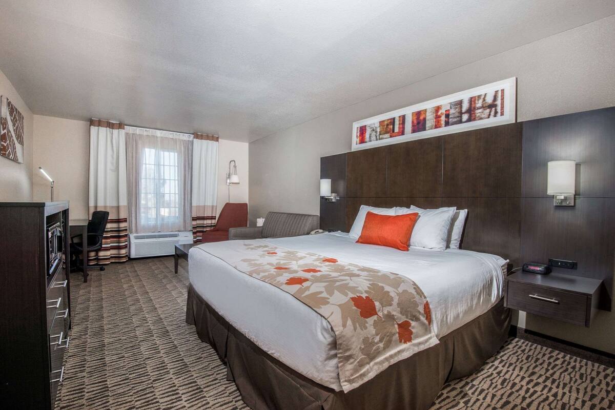 A hotel room with a large bed, a desk, a chair, a nightstand, and a wall-mounted lamp.