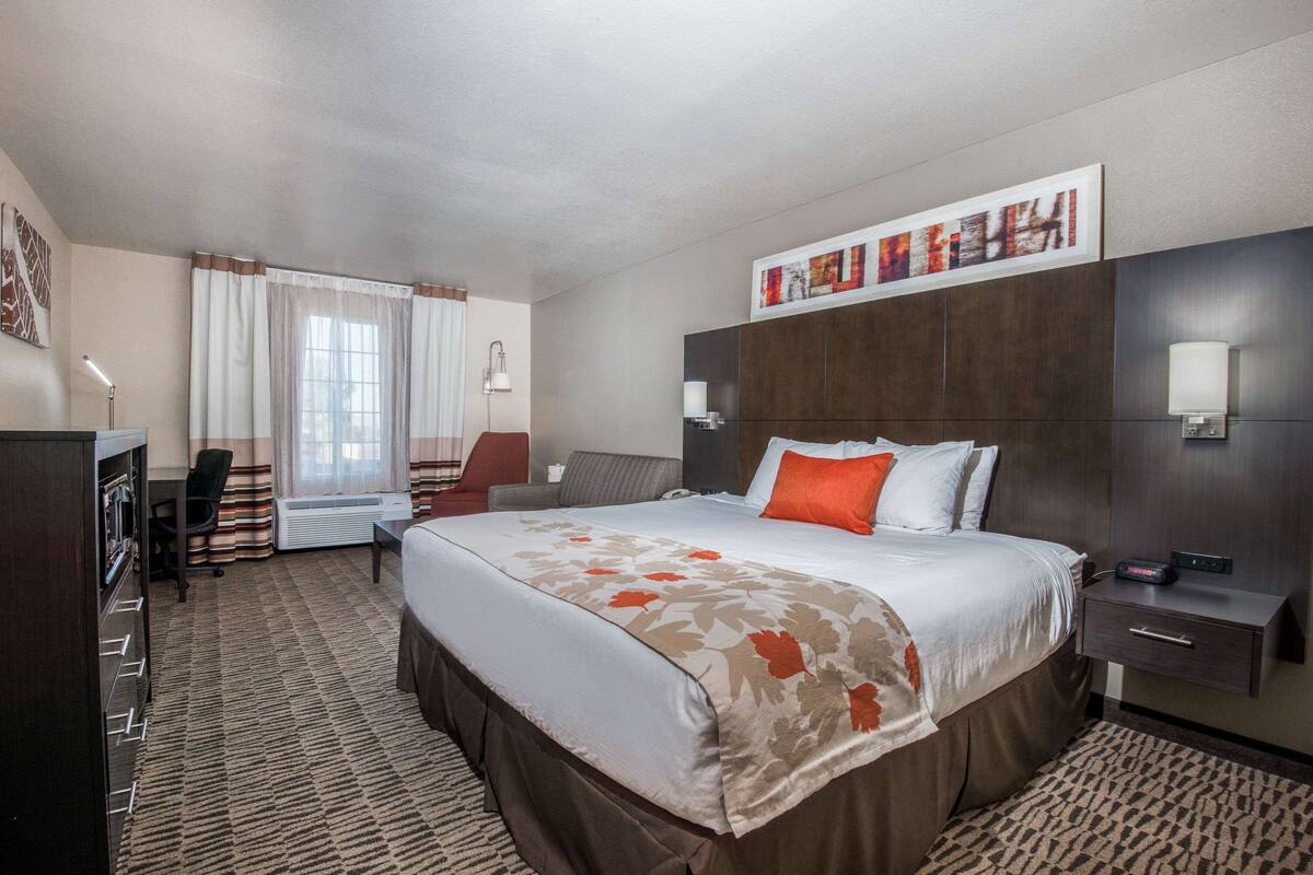 A hotel room with a large bed, a desk, a chair, a desk lamp, a wall-mounted TV, and a window with curtains.