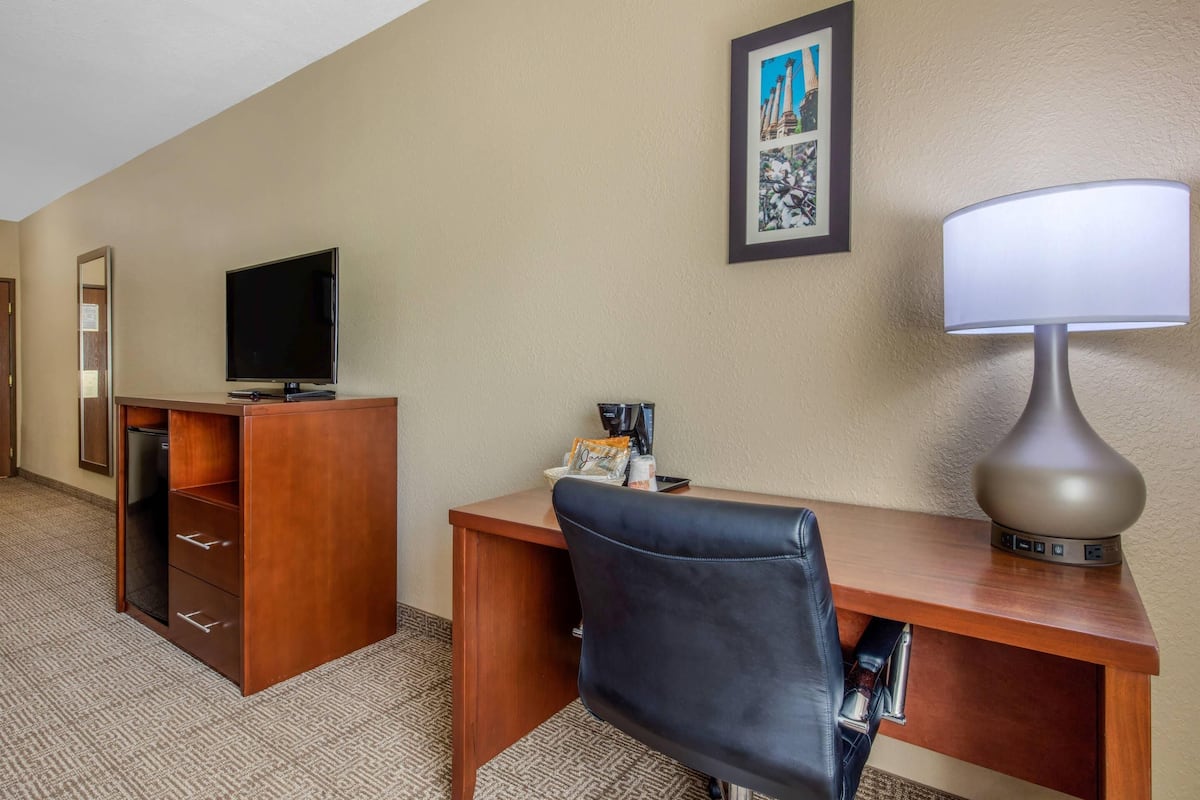 A hotel room with a wooden desk, a black leather chair, a television, a lamp, and a framed picture on the wall.