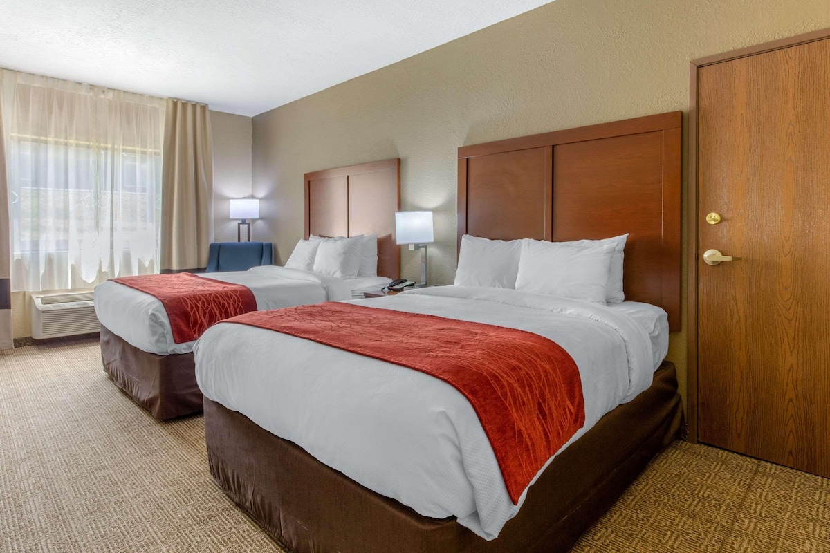 A hotel room with two beds, a desk, a chair, a lamp, and a door.