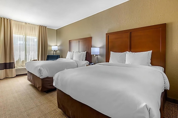 Quarto de hotel com duas camas, cada uma com cabeceira de madeira e roupa de cama branca. Há duas mesas de cabeceira com abajures, e uma janela com cortinas.