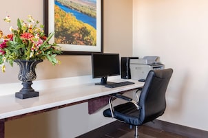 Un escritorio con computadora, impresora y silla, un florero con flores, y un cuadro de paisaje en la pared.