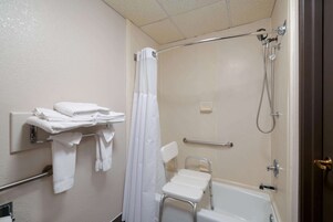 Un baño con ducha, barra de apoyo, una silla blanca y toallero.