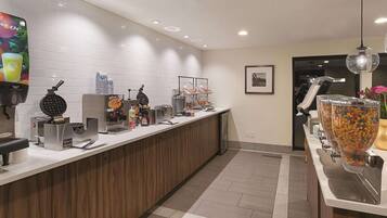 A modern hotel reception area with a wooden reception desk, a waffle maker, a coffee machine, and a variety of food items on display.