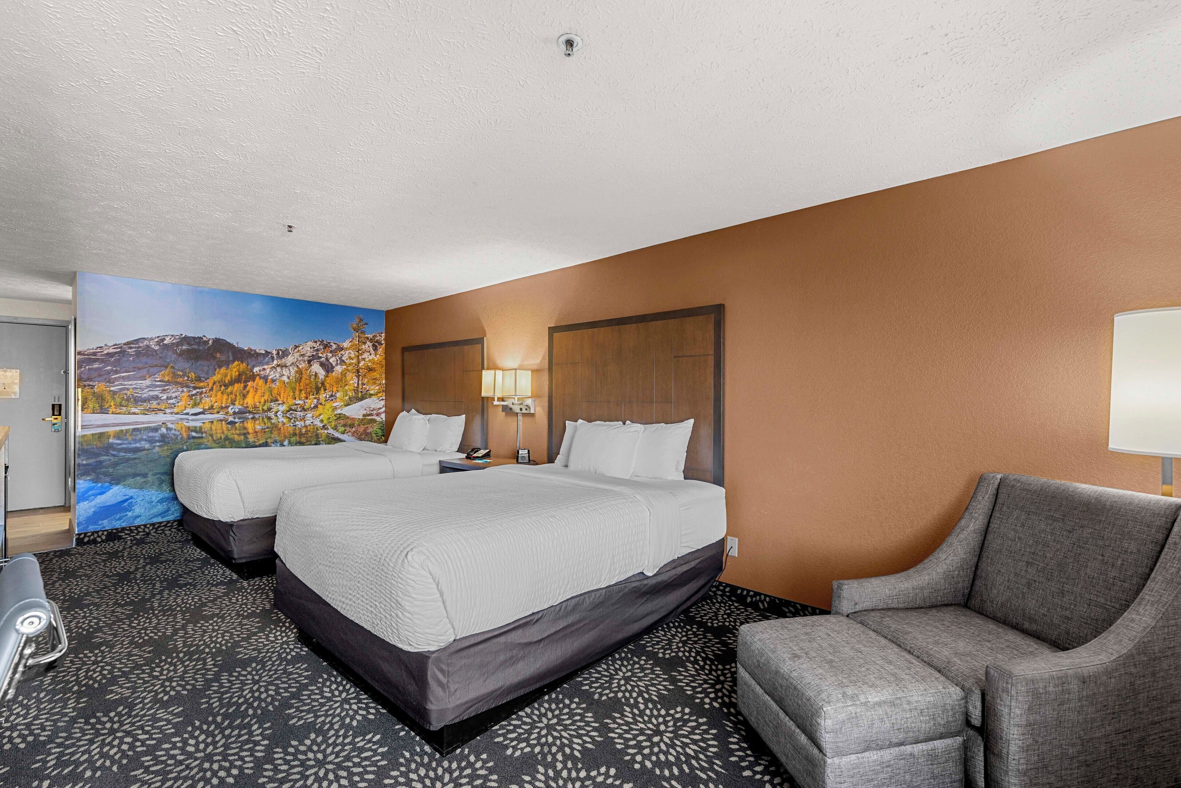 A hotel room with two beds, a gray armchair, a painting of a mountain landscape, and a large window.