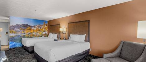 A hotel room with two beds, a gray armchair, a painting of a mountain landscape, and a large window.
