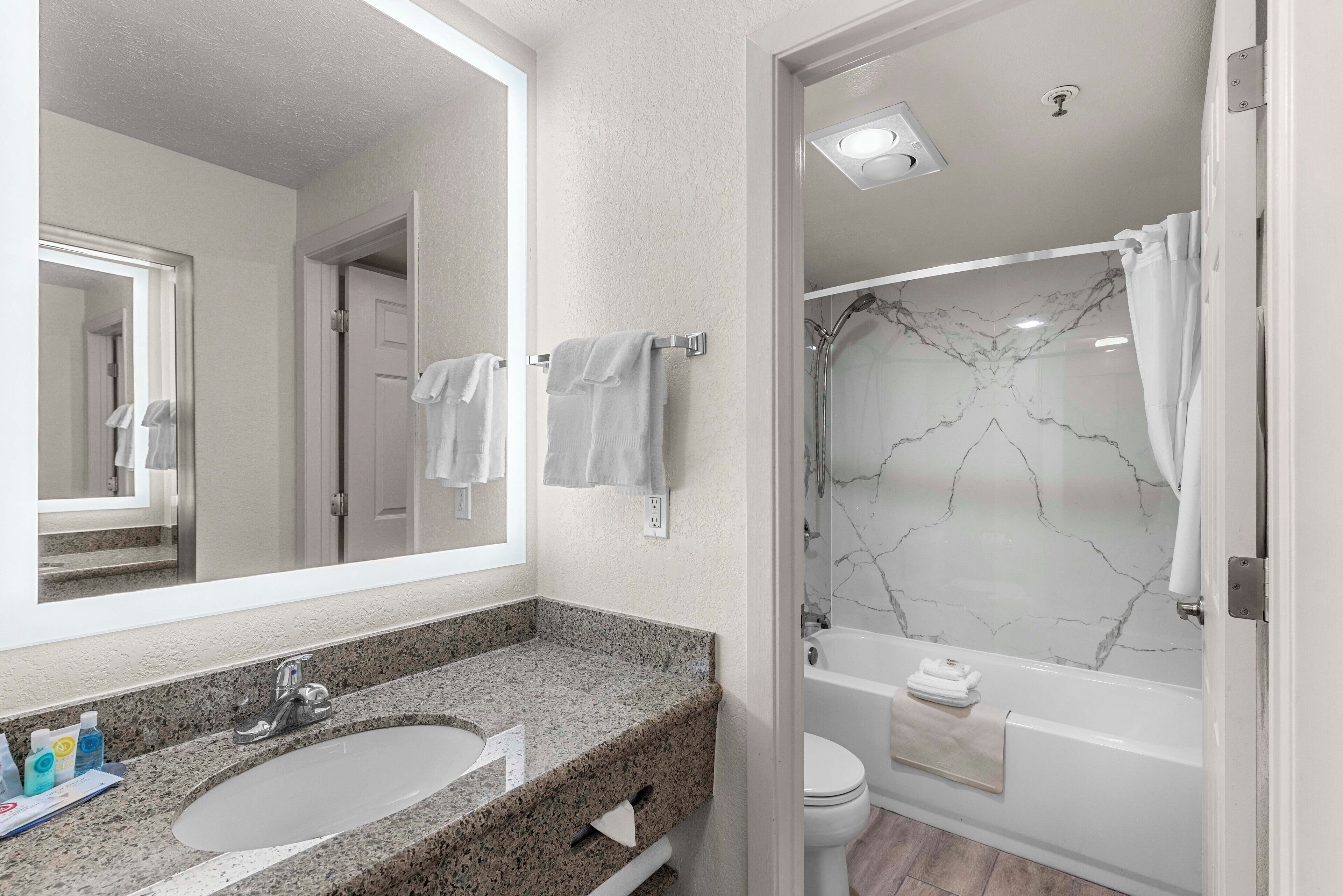 A bathroom with a granite countertop sink, a large mirror, a white toilet, and a bathtub with a marble wall.