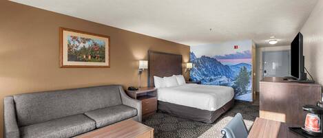 A hotel room with a sofa, a bed, a desk, and a painting of a mountain landscape.