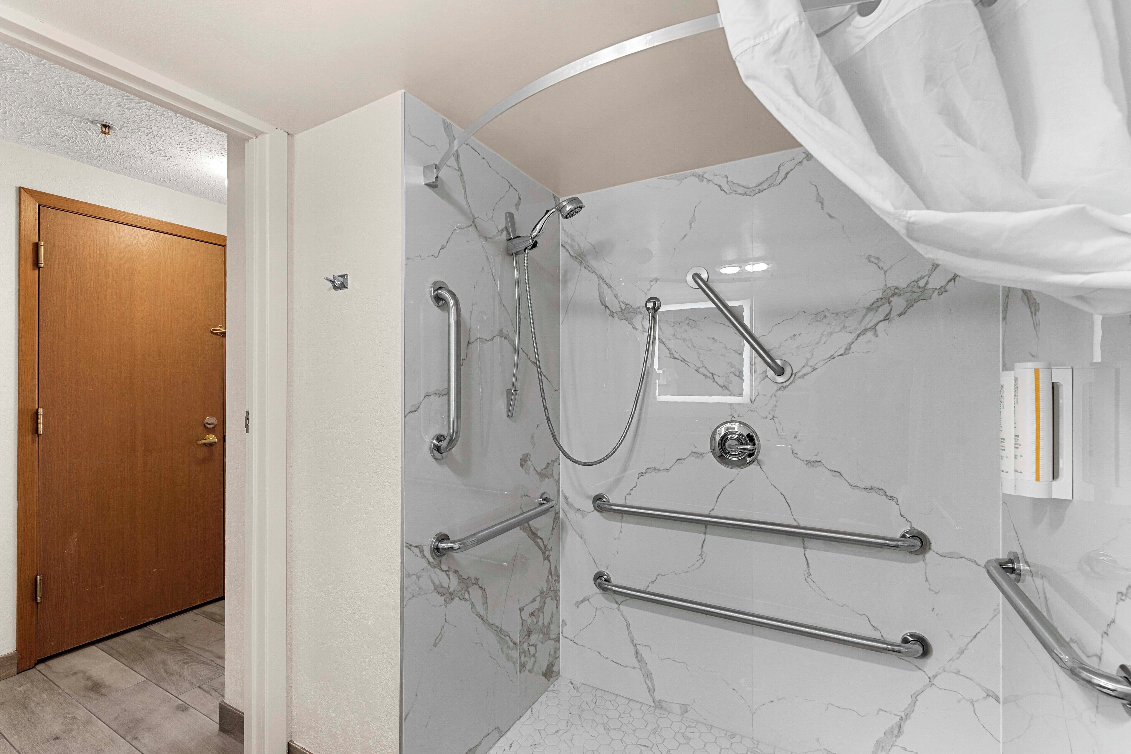 A modern bathroom with marble walls, a shower with grab bars, and a wooden door.