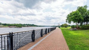 A brick pathway beside a river with a metal railing, a grassy area with a bench, and a tree. - Fireside Inn & Suites Bangor (Bangor)