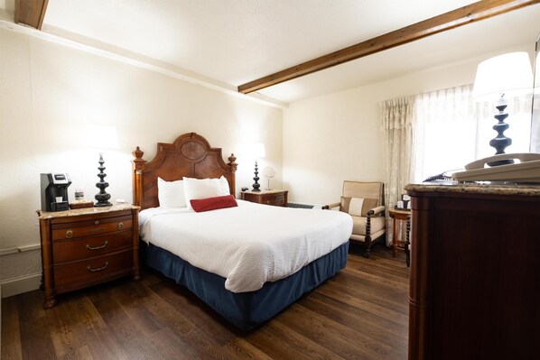 A hotel room with a wooden bed, a nightstand with a lamp, a chair, and a desk with a lamp.