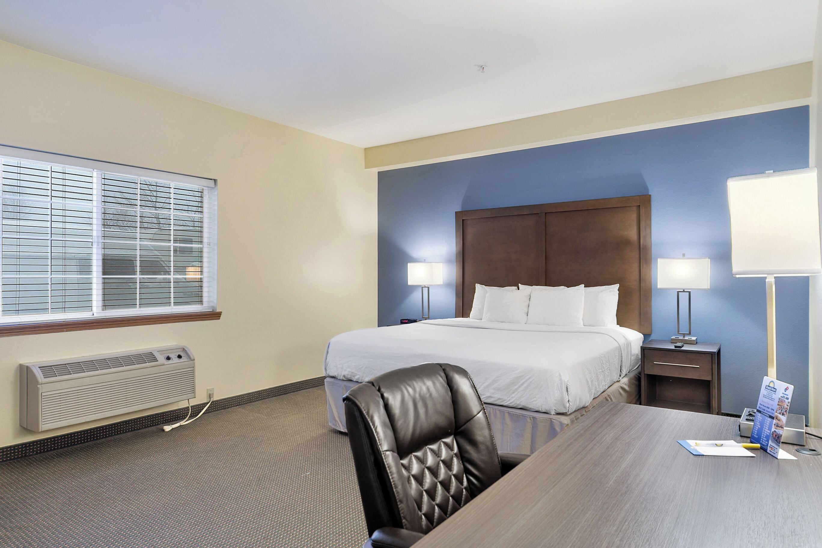 A hotel room with a bed, bedside lamps, a desk with a chair, a window with blinds, and a wall-mounted heater.