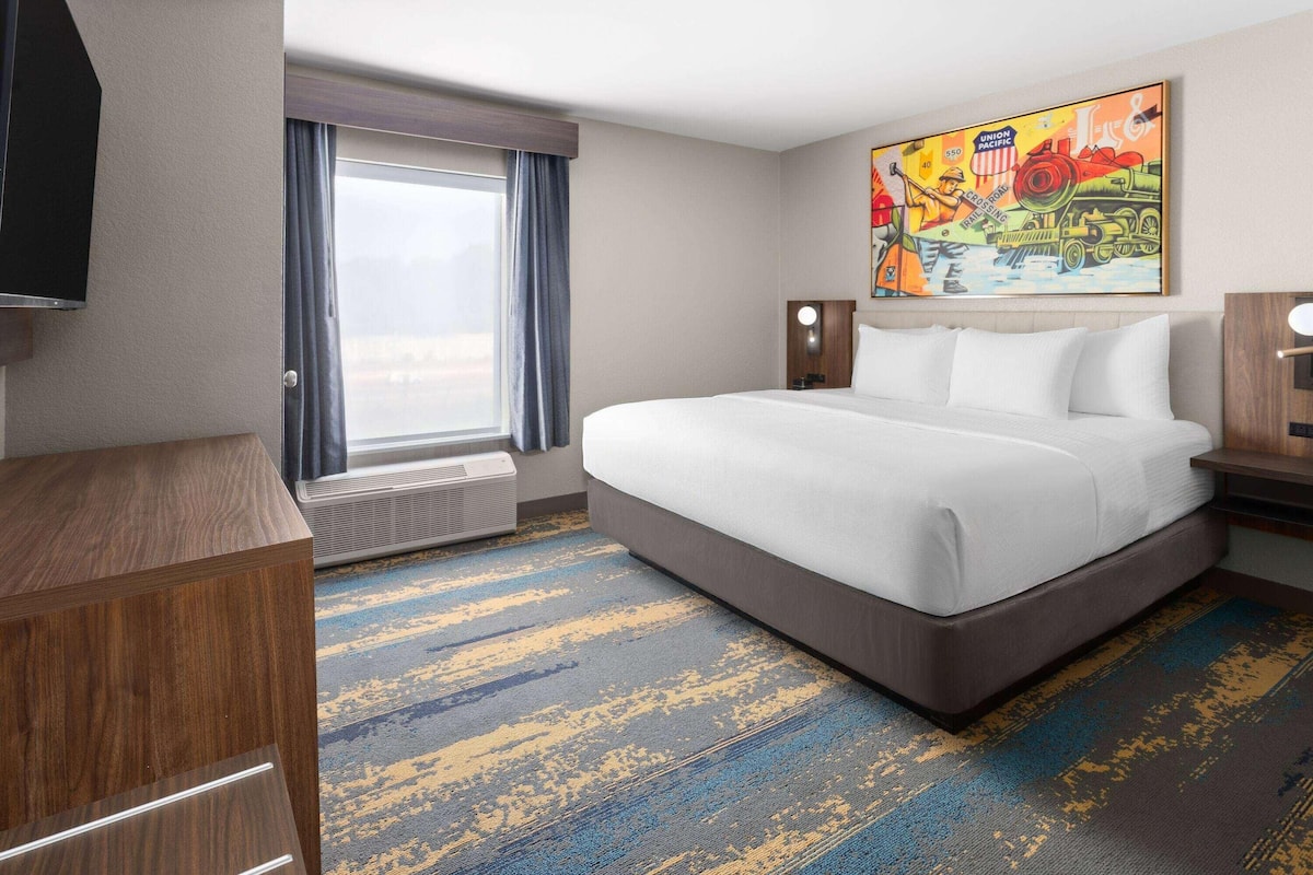 A hotel room with a large bed, a flat-screen TV, a window with curtains, a painting on the wall, and a carpeted floor.