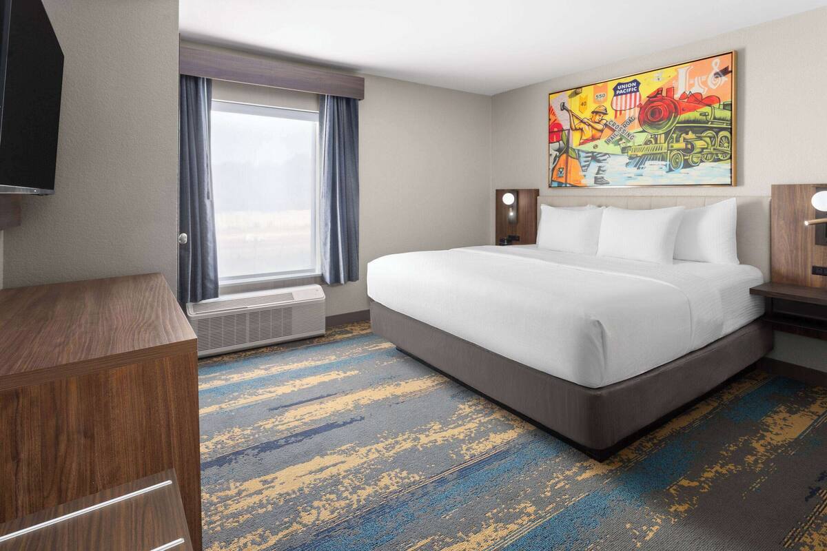 A hotel room with a large bed, a window with curtains, a painting on the wall, and a carpeted floor.