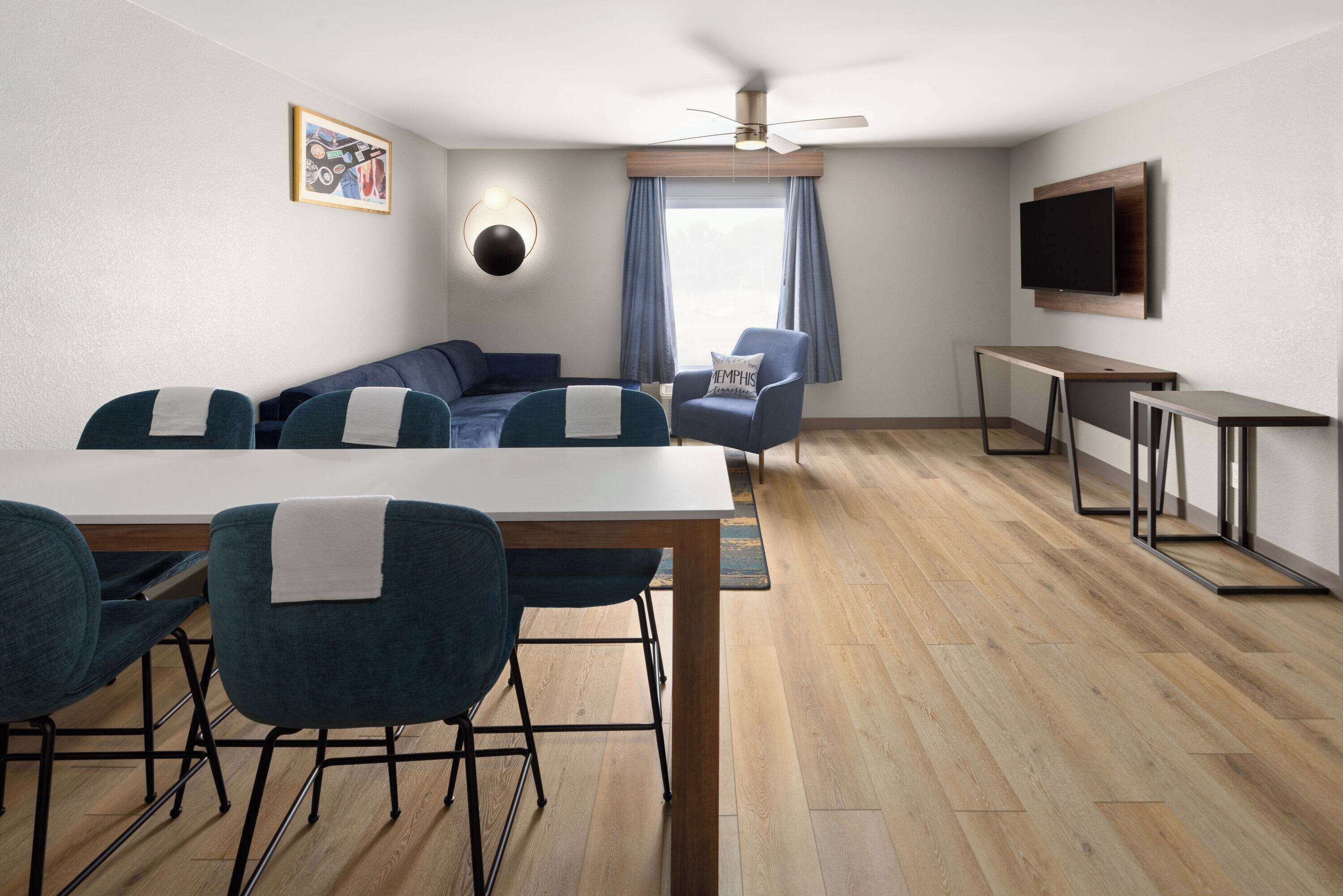 A modern hotel room with a dining area, a sofa set, a TV mounted on the wall, and a ceiling fan.