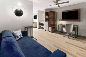Una sala moderna con un sofá azul, un televisor de pantalla plana montado en la pared, un ventilador de techo y una kitchenette al fondo.