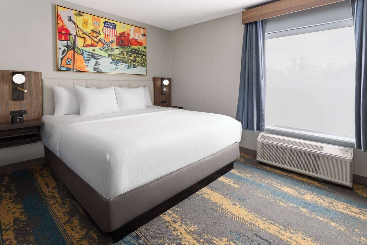 A hotel room with a large bed, a painting on the wall, a nightstand, a lamp, and a window with curtains.