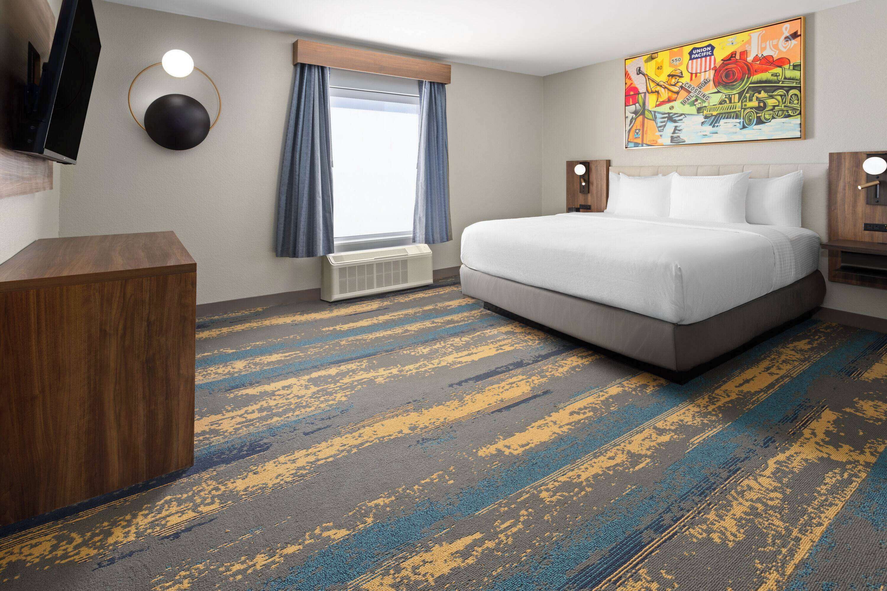 A hotel room with a large bed, a flat-screen TV mounted on the wall, a window with curtains, a radiator, and a colorful painting above the bed.