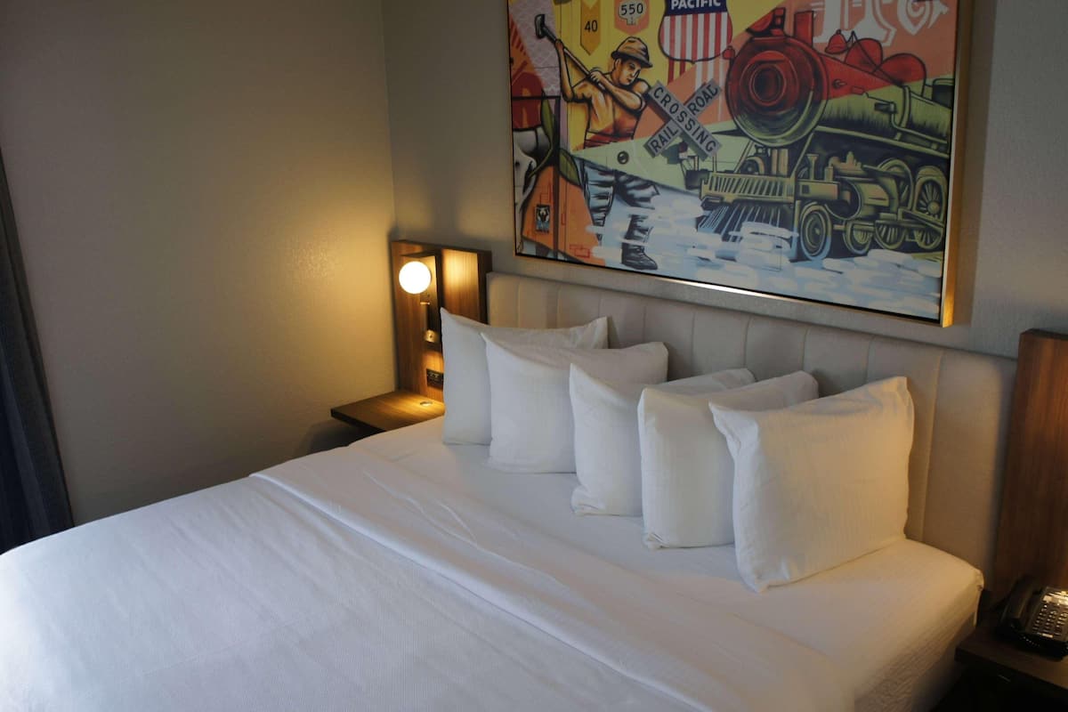 A hotel room with a neatly made bed, a nightstand with a lamp, and a wall painting of a train.
