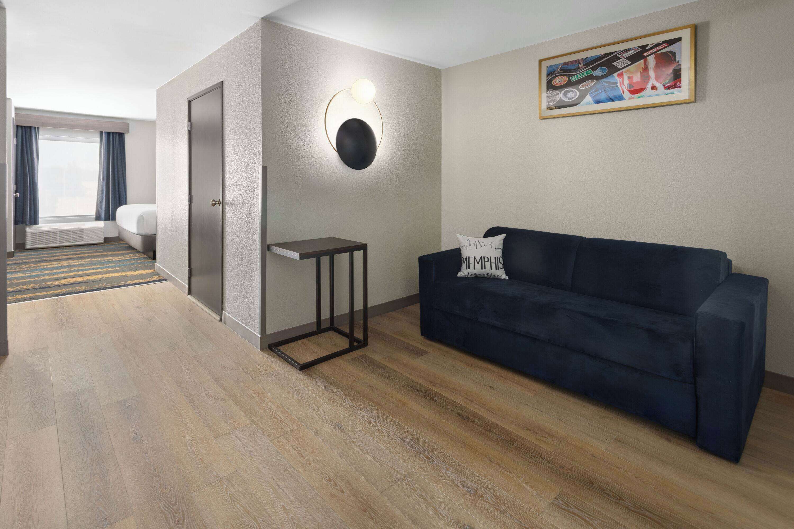A modern hotel room with a dark blue sofa, a small side table, a round wall light, and a painting on the wall.