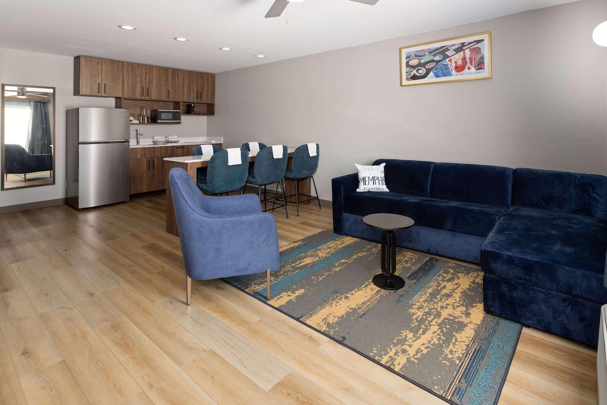 Ein modernes Wohnzimmer mit einem dunkelblauen Sofa, einem blauen Sessel und einem kleinen runden Tisch. Es gibt eine Küchenzeile mit Holzschränken und einem Kühlschrank.