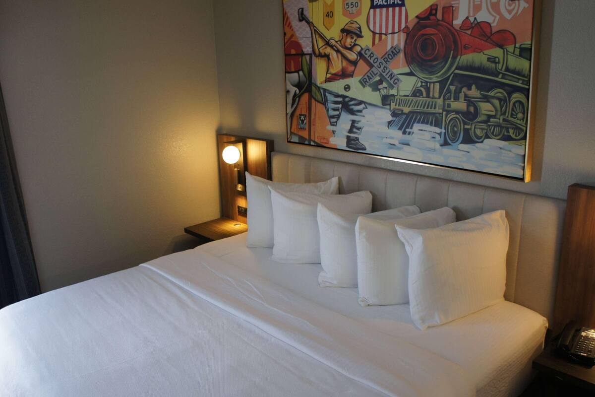 A hotel room with a neatly made bed, a nightstand with a lamp, and a colorful painting on the wall depicting a train scene.