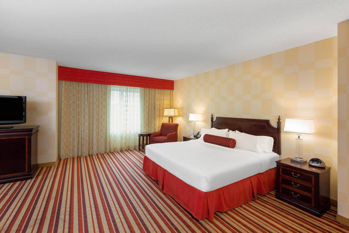 A hotel room with a large bed, two bedside tables with lamps, a TV, a chair, and a window with curtains.