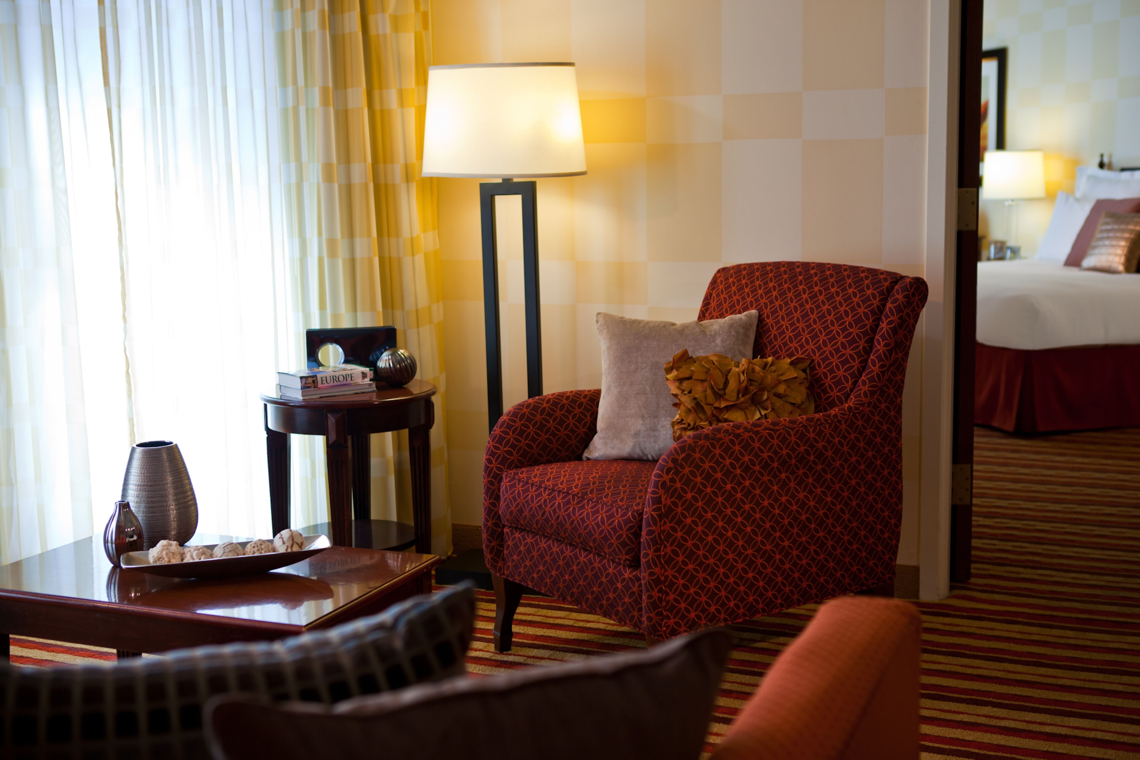 A hotel room with a patterned armchair, a bedside table with a lamp, and a view into another room with a bed.