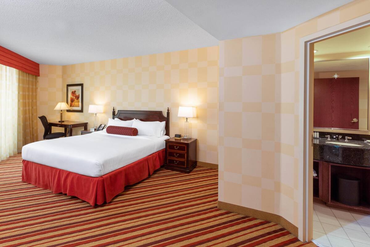A hotel room with a large bed, two bedside tables with lamps, a desk with a chair, and a bathroom visible through an open door.