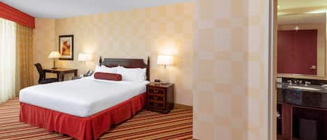 Una habitación de hotel con una cama grande, dos mesitas de noche con lámparas, un escritorio con una silla y un baño visible a través de una puerta abierta.