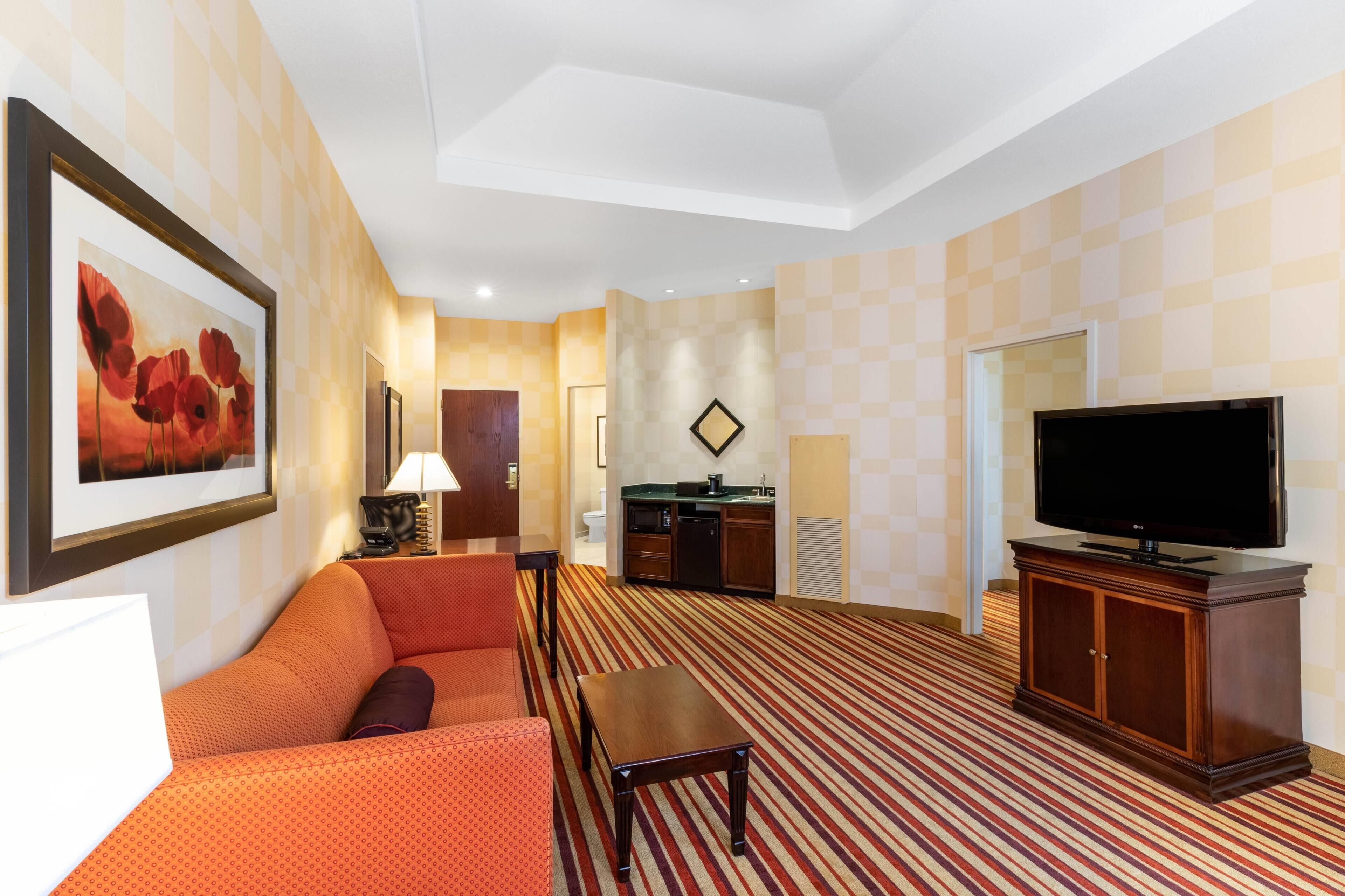 A hotel room with a sofa, a TV, a small table, a kitchen area, and a framed picture on the wall.