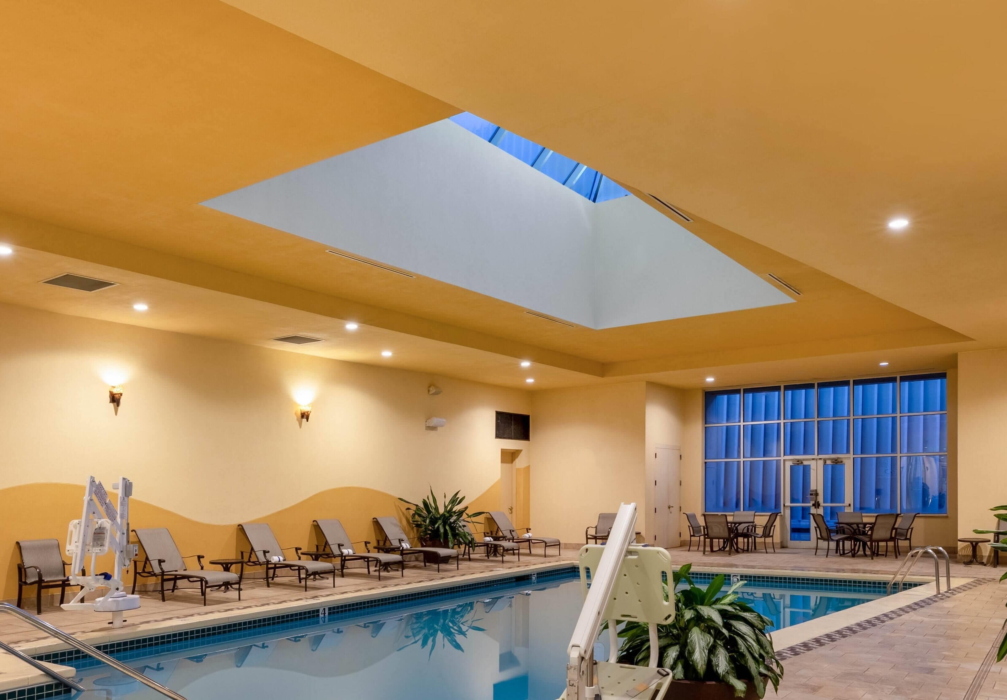 Piscina interna iluminada com luzes no teto, claraboias e espreguiçadeiras.