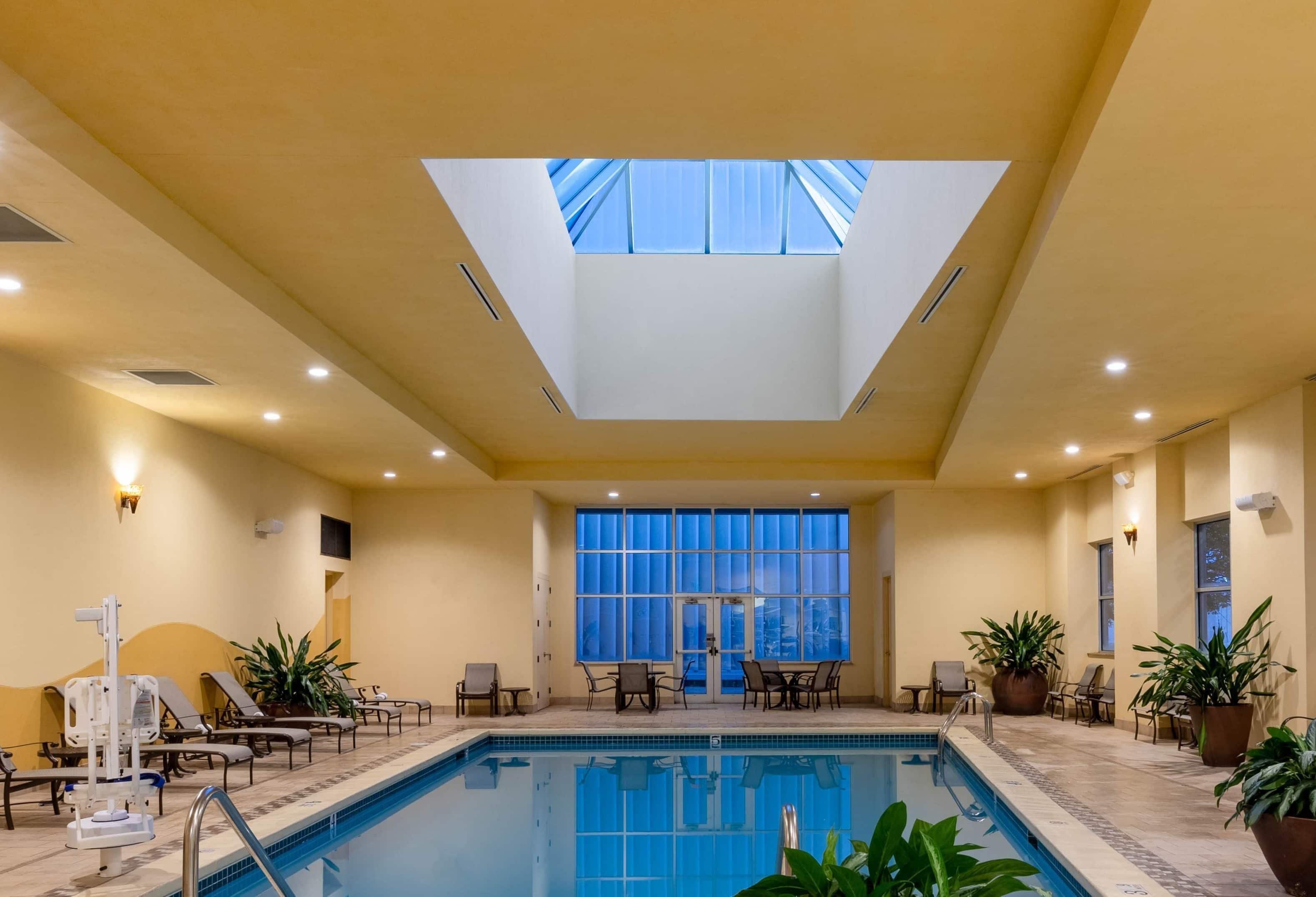 An indoor swimming pool with a skylight, lounge chairs, and potted plants.