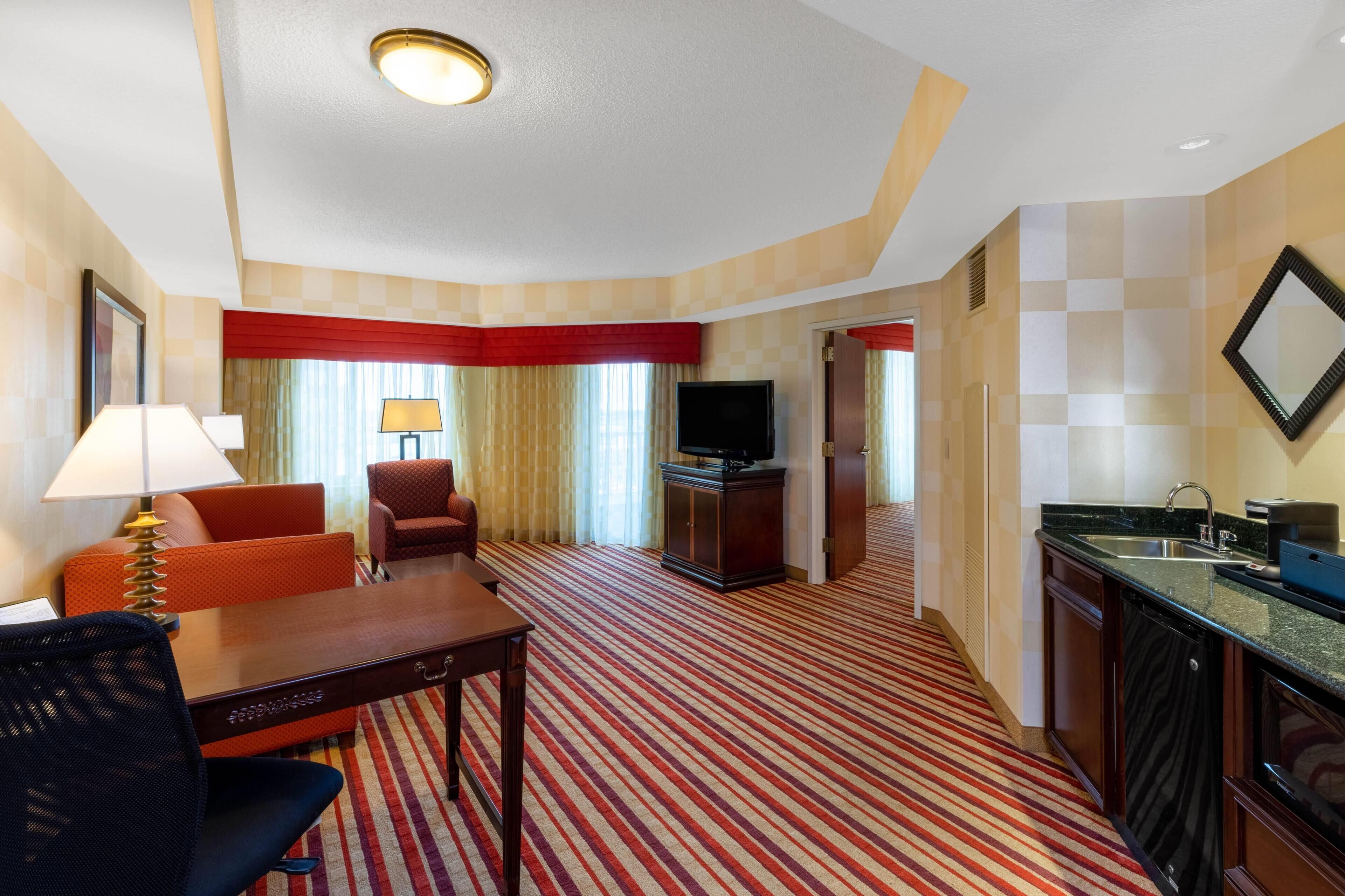 A hotel room with a dining table, two chairs, a sofa, a TV, a kitchen area with a sink, and a large window with curtains.