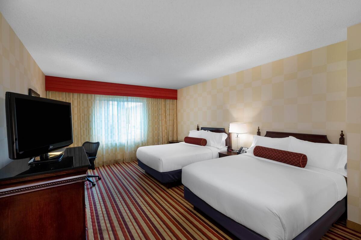 A hotel room with two beds, a TV, a desk, and a window with curtains.