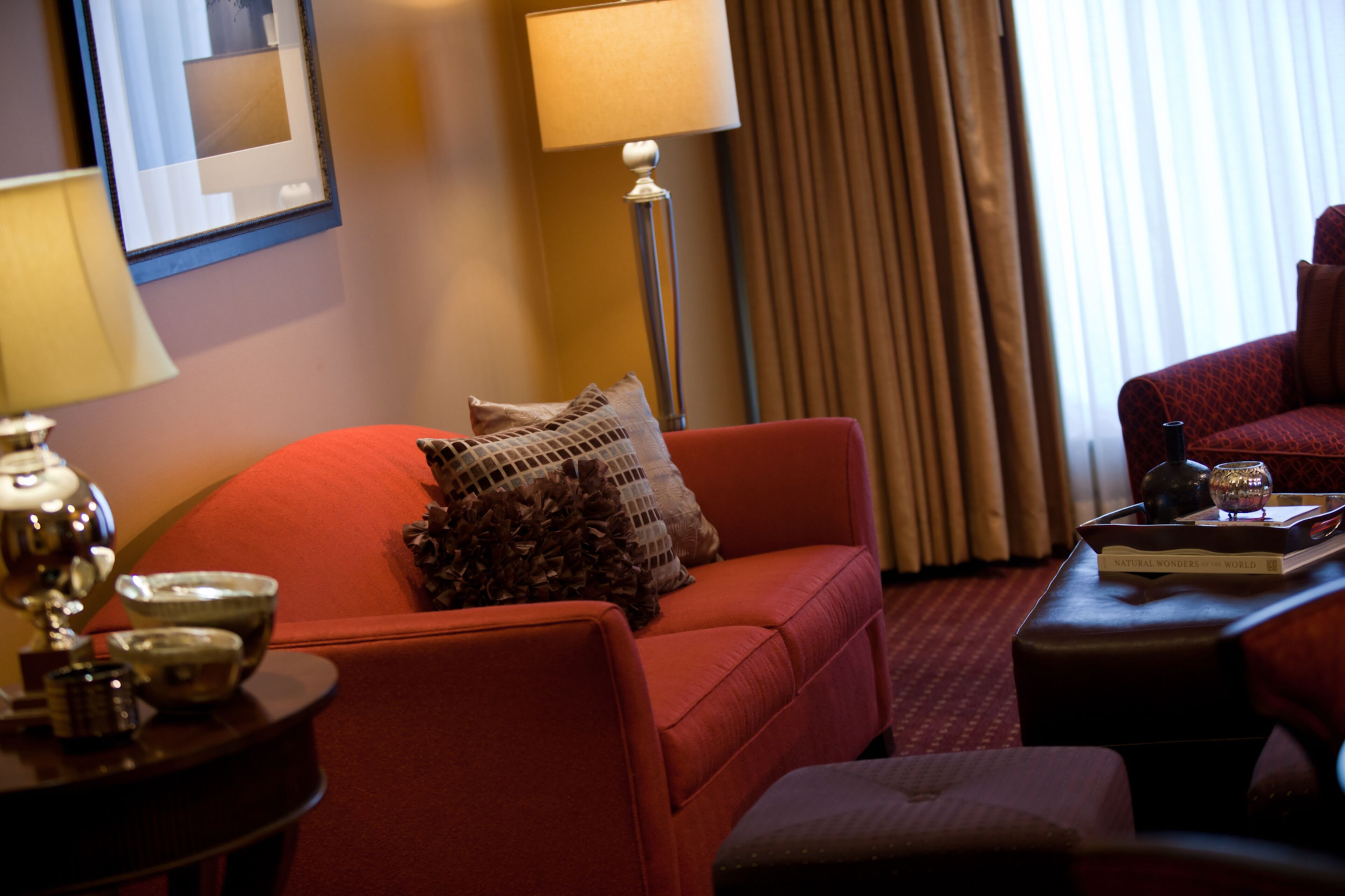 A hotel room with a red sofa, a side table with a lamp, a tray with a book and a glass, and a window with curtains.