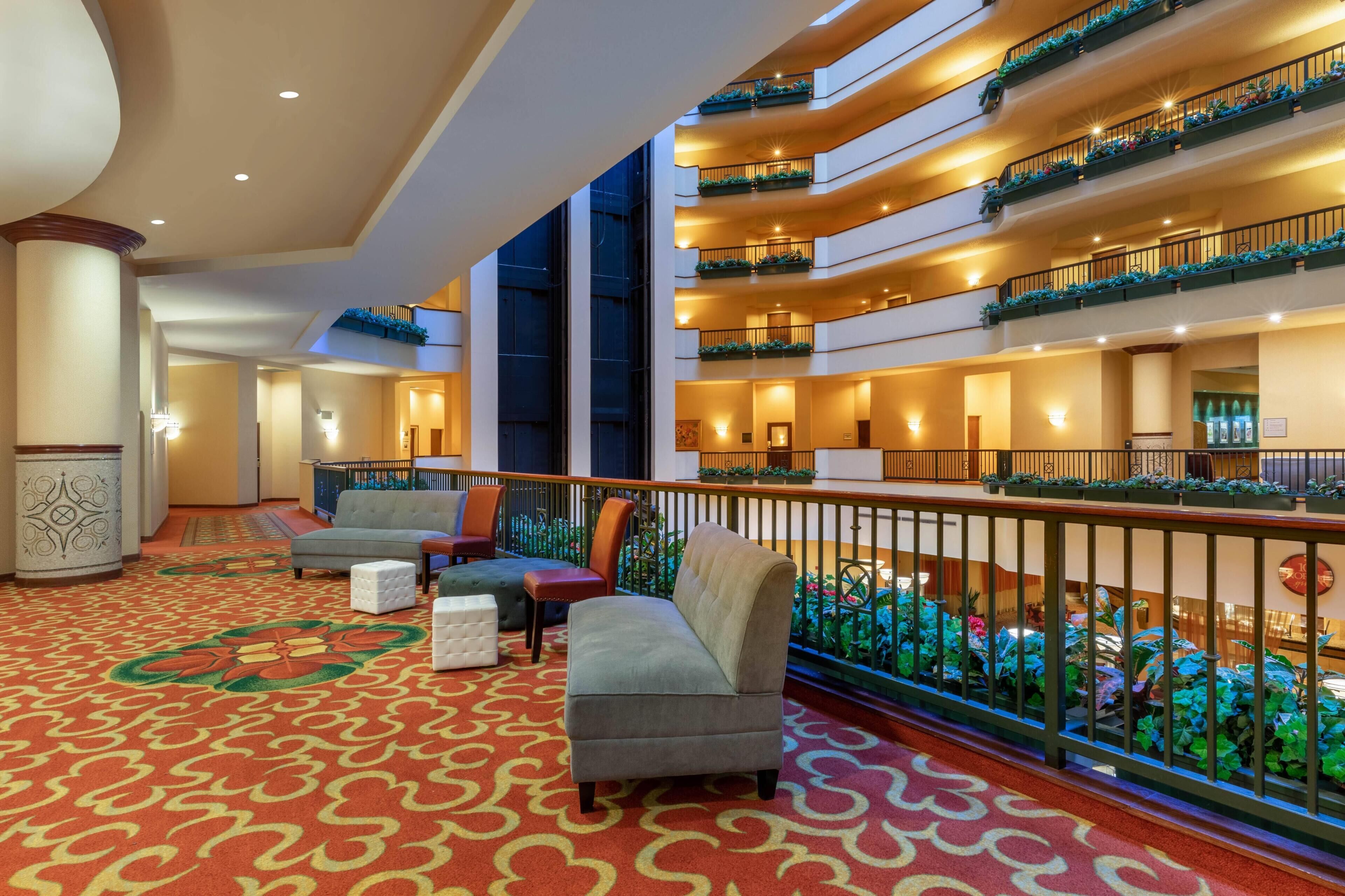 A hotel lobby with a red patterned carpet, multiple seating areas, and balconies with plants.