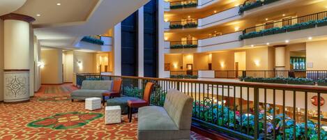 A hotel lobby with a red patterned carpet, multiple seating areas, and balconies with plants.